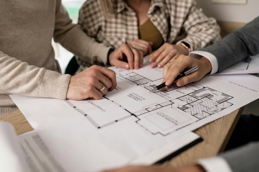 Three people reviewing a residential floor plan together at a table, pointing to architectural drawings during a home design or real estate meeting.